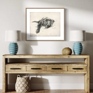 May include: A black and white drawing of a dog lying down on a bed, framed in a light wood frame and hanging on a white wall above a wooden console table with two lamps and a woven basket.