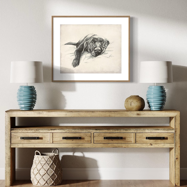 May include: A black and white drawing of a dog lying down on a bed, framed in a light wood frame and hanging on a white wall above a wooden console table with two lamps and a woven basket.