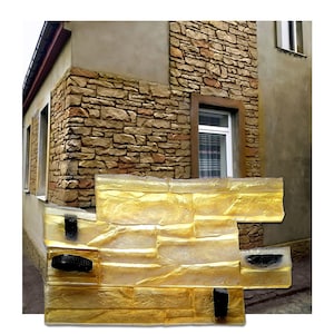 Può includere: Uno stampo in silicone color oro per creare una texture da muro in pietra. Lo stampo è composto da tre sezioni e presenta una superficie testurizzata che ricorda un muro di mattoni. Lo stampo è mostrato su uno sfondo di un vero muro in pietra.