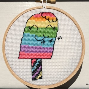 May include: A rainbow popsicle cross-stitch pattern in a wooden embroidery hoop. The popsicle is smiling and has a striped stick.