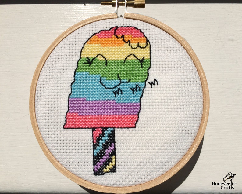 May include: A rainbow popsicle cross-stitch pattern in a wooden embroidery hoop. The popsicle is smiling and has a striped stick.