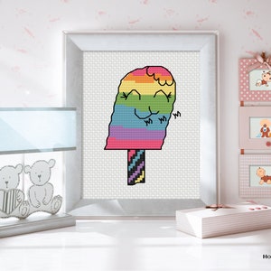 May include: Cross stitch pattern of a rainbow popsicle with a smiling face. The popsicle is on a stick with a striped design. The pattern is framed and displayed on a white wall.