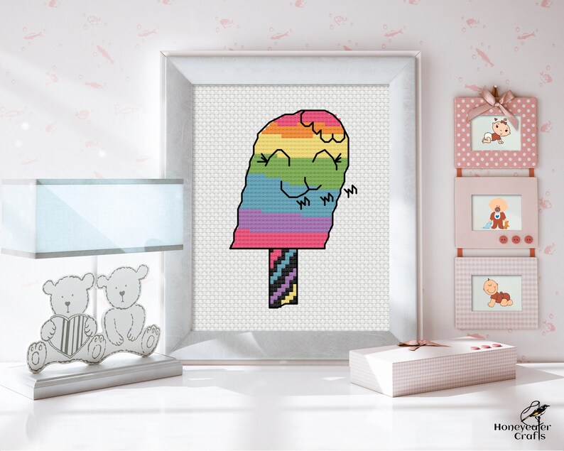 May include: Cross stitch pattern of a rainbow popsicle with a smiling face. The popsicle is on a stick with a striped design. The pattern is framed and displayed on a white wall.