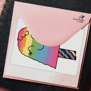 May include: A rainbow popsicle cross stitch pattern with a black and white striped stick. The pattern is on white Aida cloth and is framed in a white frame. The pattern is from Honeyweaver Crafts.