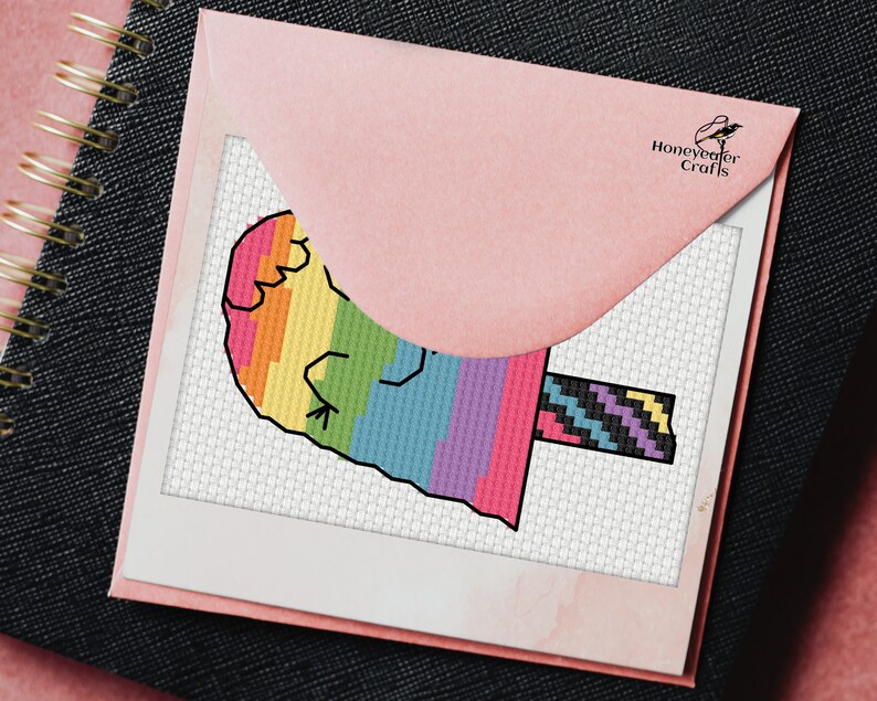 May include: A rainbow popsicle cross stitch pattern with a black and white striped stick. The pattern is on white Aida cloth and is framed in a white frame. The pattern is from Honeyweaver Crafts.