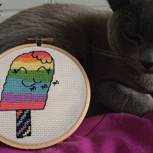 May include: A rainbow popsicle cross-stitch pattern in a wooden embroidery hoop. The popsicle has a smiling face and is surrounded by a white background.
