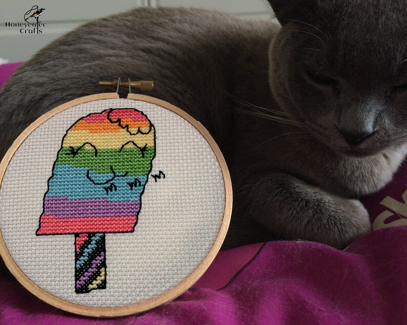 May include: A rainbow popsicle cross-stitch pattern in a wooden embroidery hoop. The popsicle has a smiling face and is surrounded by a white background.
