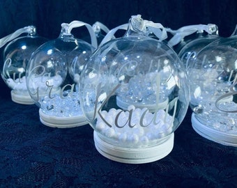 May include: Five clear glass baubles with white bases and white glitter inside. Each bauble has a different name written on it in white lettering: "Clara", "Gra", "Kaut", "Izzy", and "Eliza".