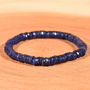 Könnte beinhalten: Ein dunkelblaues Edelsteinarmband. Das Armband besteht aus kleinen, facettierten, runden Edelsteinen, die aneinandergereiht sind. Die Edelsteine haben eine tiefe, satte blaue Farbe. Das Armband wird auf einer hellbraunen Oberfläche präsentiert.