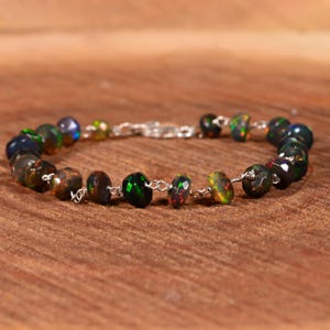 以下が含まれることがあります： 緑、赤、青の輝きを持つ、黒オパールのファセットビーズのブレスレット。ビーズは繊細なシルバーチェーンに通されています。ブレスレットは木製の表面に展示されています。