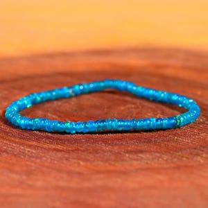 Puede incluir: Una delicada pulsera de cuentas azules. La pulsera está hecha de pequeñas cuentas redondas en diferentes tonos de azul, creando un efecto brillante. La pulsera se muestra sobre una superficie de madera.