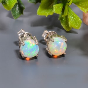 Könnte beinhalten: Ein Paar silberne Ohrstecker mit runden Opal-Edelsteinen. Die Opale haben eine weiße Basis mit irisierenden Schimmern in Grün, Blau und Orange.