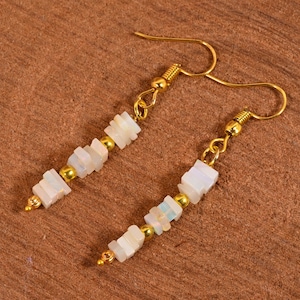 Può includere: Un paio di orecchini pendenti con ganci e dettagli color oro. Ogni orecchino presenta piccole perline rettangolari bianche e perline color oro. Gli orecchini sono esposti su una superficie di legno marrone.