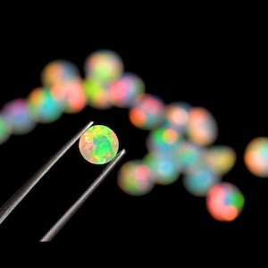 May include: A small, iridescent opal gemstone held by tweezers against a black background. The opal is a pale green color with flashes of pink, blue, and yellow.