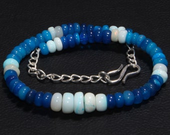 May include: A beaded bracelet featuring round, disc-shaped beads in various shades of blue, ranging from light sky blue to deep navy, interspersed with opaque white beads. The bracelet includes a silver-toned metal chain and a hook-style clasp.