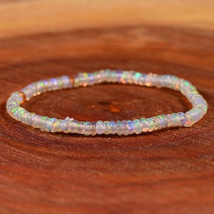 May include: A delicate bracelet composed of small, iridescent opal beads. The beads display a range of colors, including white, blue, green, and orange, and are strung together to form a flexible band. The bracelet is displayed on a wooden surface.