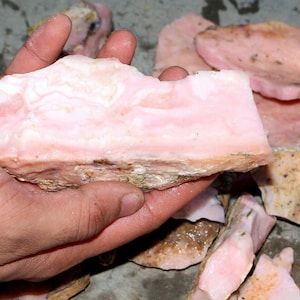 May include: A large piece of pink opal with a rough, natural texture. The opal is translucent and has a light pink colour. The opal is being held in a hand.