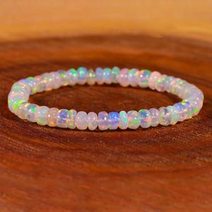 Puede incluir: Un brazalete de cuentas de ópalo blanco con colores arcoíris iridiscentes. El brazalete está hecho de pequeñas cuentas redondas y está enhebrado en un cordón elástico transparente.