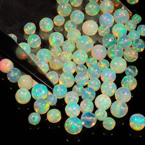 Op de afbeelding: Een verzameling van regenboogkleurige opaal kralen in verschillende tinten groen, blauw en wit. De kralen zijn rond en glad en zijn verspreid over een zwart oppervlak.