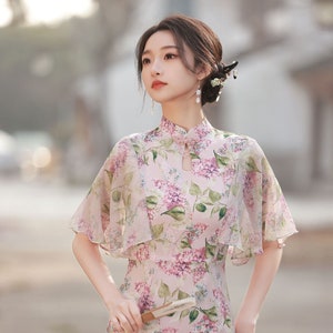 May include: A woman wearing a pink floral dress with a mandarin collar and short sleeves. The dress has a delicate floral pattern and a flowing skirt. She is holding a small, decorative fan in her hand.