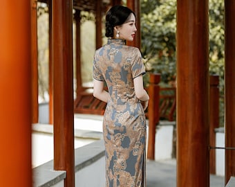May include: A woman wearing a long, blue and gold cheongsam dress with a floral pattern. The dress has a mandarin collar and short sleeves. She is standing in a garden setting.