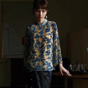 Pode incluir: Uma mulher vestindo uma blusa de estampa floral amarela e azul com colarinho mandarim e uma saia preta. Ela está em pé em frente a um fundo escuro.