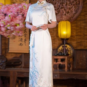 May include: A white dress with a mandarin collar and short sleeves. The dress has a floral pattern in blue and pink. The dress is worn by a person standing in a room with a wooden floor and a traditional Chinese decor.