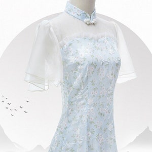 May include: A light blue dress with a white floral pattern and sheer white sleeves. The dress has a mandarin collar and a button closure at the neck.