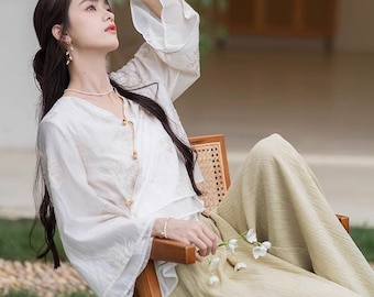 May include: A woman wearing a white blouse with a button closure and long sleeves sits on a wooden chair. She is wearing light green pants and has long black hair. She is holding a small bouquet of white flowers.