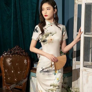 May include: A white cheongsam dress with a floral print of lotus flowers and leaves. The dress has a mandarin collar and short sleeves. The model is holding a wooden fan.
