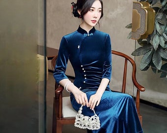 May include: A long, dark teal velvet dress with a mandarin collar and three-quarter sleeves. The dress features a row of pearl buttons down the front and a slight ruched detail at the waist. A small, beaded purse is held in the hand.