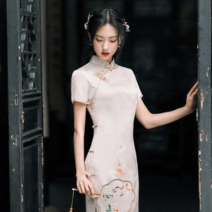 May include: A woman in a white cheongsam dress with floral embroidery stands in a doorway. She is holding a white fan with floral embroidery and a tassel.