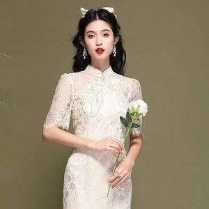 May include: A white lace dress with a mandarin collar and short sleeves. The dress has a floral pattern and is fitted to the body. The dress is knee-length and has a lace trim at the hem.