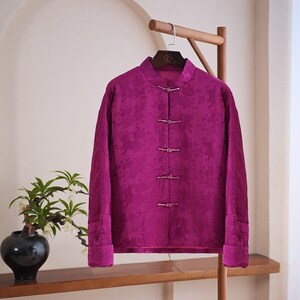 Peut inclure: Une veste en soie magenta avec un col mandarin et cinq boutons décoratifs. La veste est suspendue à un cintre en bois.