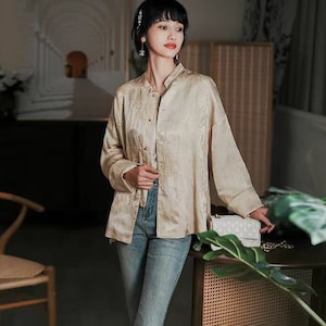 Könnte beinhalten: Eine Frau trägt ein hellbeiges, langärmeliges Hemd mit Stehkragen und einem Blumenmuster. Sie trägt auch eine blaue Jeans und weiße Turnschuhe.