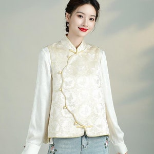 May include: A white sleeveless top with a gold embroidered pattern and a mandarin collar. The top has a diagonal closure with gold buttons.