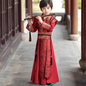 May include: A young person wearing a red and gold traditional Chinese robe with black trim and a long black tassel. They are holding a wooden flute and playing it.