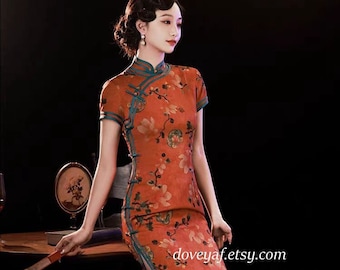 May include: A woman wearing a red cheongsam with a floral print and teal trim. The cheongsam has a mandarin collar and is fitted to the body. The woman is sitting on a wooden table with a book and a fan.