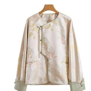 May include: A beige silk jacket with a floral and geometric pattern. The jacket has a mandarin collar, long sleeves, and a tie closure. The sleeves have green trim.