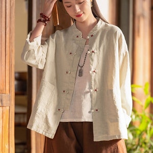 May include: A light beige linen shirt with a mandarin collar and red buttons. The shirt has a pocket on the left side and is worn over a white undershirt. The person is wearing brown trousers.
