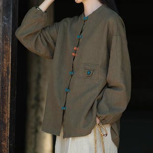 Può includere: Camicia verde oliva a maniche lunghe con colletto alla coreana e una piccola tasca. La camicia presenta bottoni decorativi nei toni dell'arancione e del turchese. La modella indossa una gonna beige e tiene una collana di perline.