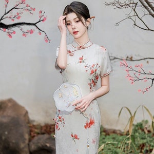 May include: A woman wearing a white dress with a floral pattern and a red trim. The dress has a mandarin collar and short sleeves. The woman is holding a white fan with a floral design. The text on the image reads "东方神韵" which translates to "Oriental Charm".