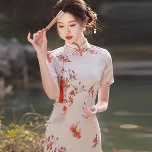 May include: A white cheongsam dress with a floral print and red accents. The dress has a mandarin collar and short sleeves. The dress is fitted and has a side slit.