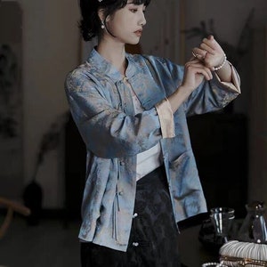 May include: A woman wearing a light blue patterned jacket with a mandarin collar and a black skirt with buttons. She is wearing a pearl bracelet and pearl earrings.