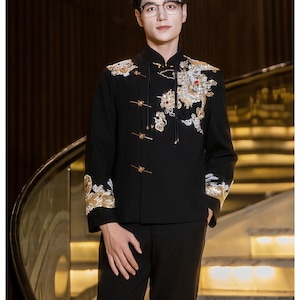 May include: Black formal jacket with ornate gold and white embroidery, featuring a dragon design on the front and sleeves. The jacket has a mandarin collar and gold frog closures. The person is wearing black pants and glasses.
