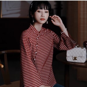 Pode incluir: Uma blusa xadrez vermelha e branca com gola mandarim e mangas compridas. A blusa tem fecho de botões e é feita de um tecido leve.