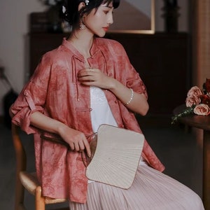 May include: A woman wearing a pink, loose-fitting,  button-up shirt with a white under shirt. She is holding a white fan and wearing a light pink pleated skirt.