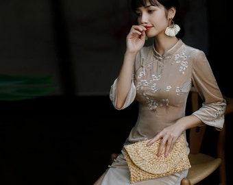 May include: A beige velvet dress with floral embroidery. The dress has a mandarin collar, long sleeves, and a side slit. A woven clutch is held in the lap. The dress is a traditional style.
