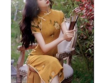 May include: A woman wearing a yellow floral print cheongsam dress with a mandarin collar and short sleeves. She is holding a wooden fan and sitting on a wooden chair in a garden setting.