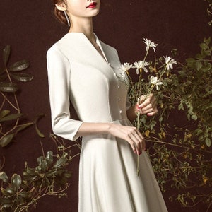 Może przedstawiać: Biała sukienka z dekoltem w kształcie litery V i długimi rękawami. Sukienka ma dopasowany gorset i rozkloszowaną spódnicę. Modelka trzyma bukiet białych kwiatów.